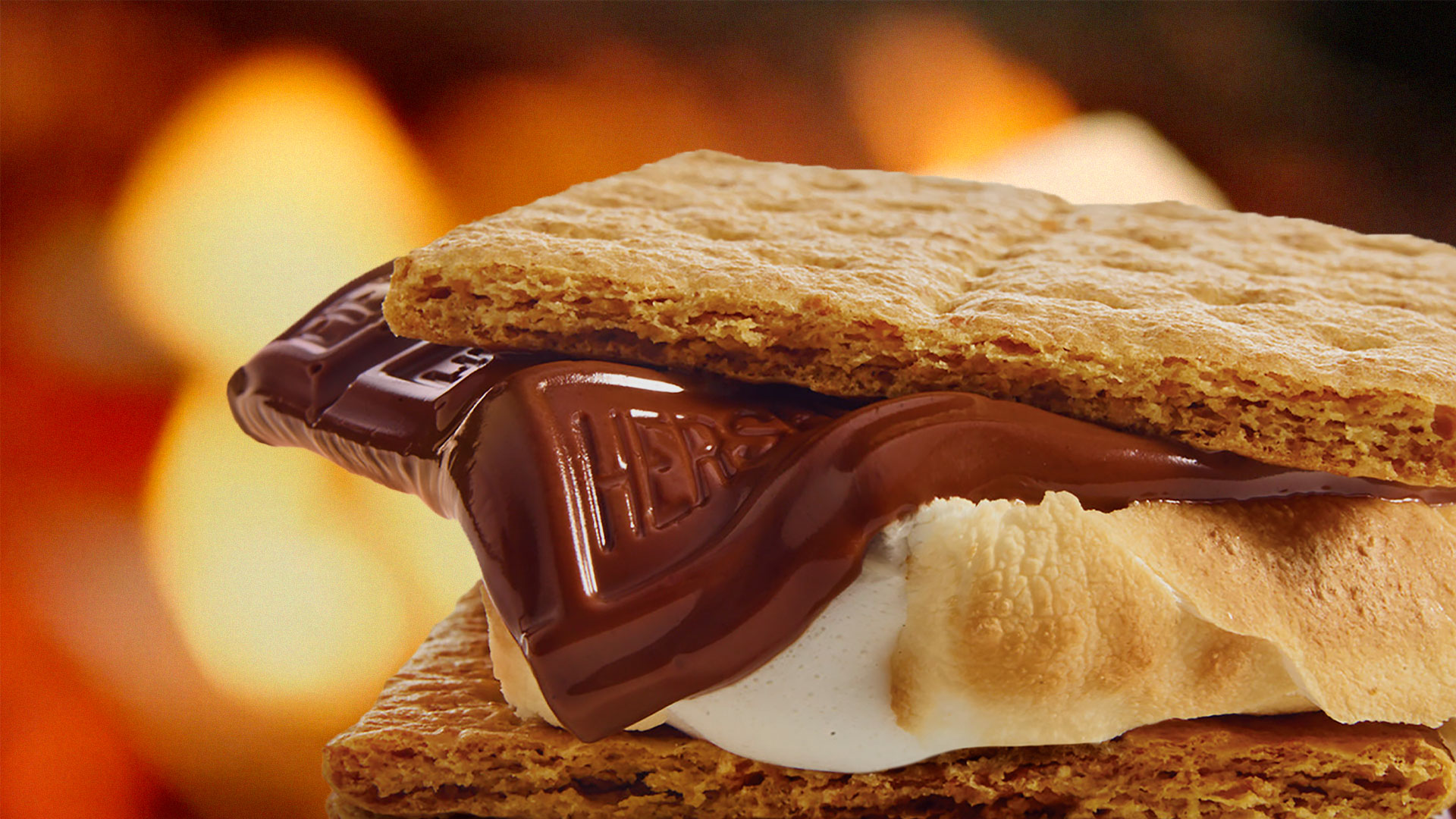 Mouth-watering photo of Hershey's chocolate melting under graham cracker and over a toasted marshmallow. A giant fire is behind the S'mores